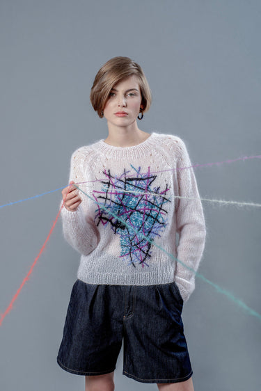 Woman wearing light pink mohair sweater and holding wool yarns going towards camera. The sweater has colourful embroidered lines forming an abstract shape in the center.